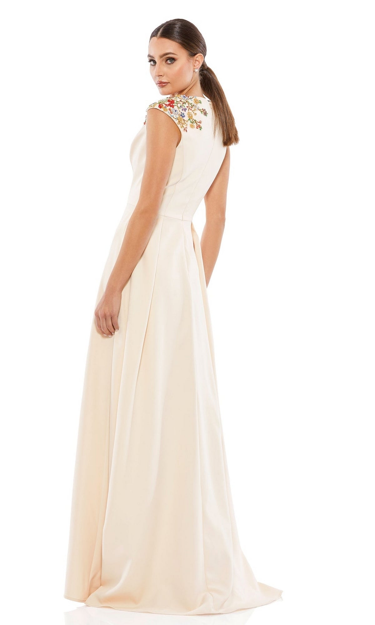 Woman wears the Mac Duggal 26540 cap-sleeve long formal dress, featuring cream-colored fabric with embroidered floral embellishments on the shoulders, as she stands and looks over her shoulder against a white background.